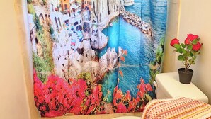 A bathroom with a colorful shower curtain depicting a coastal town, a white toilet, a pink bath mat, and a small potted plant.