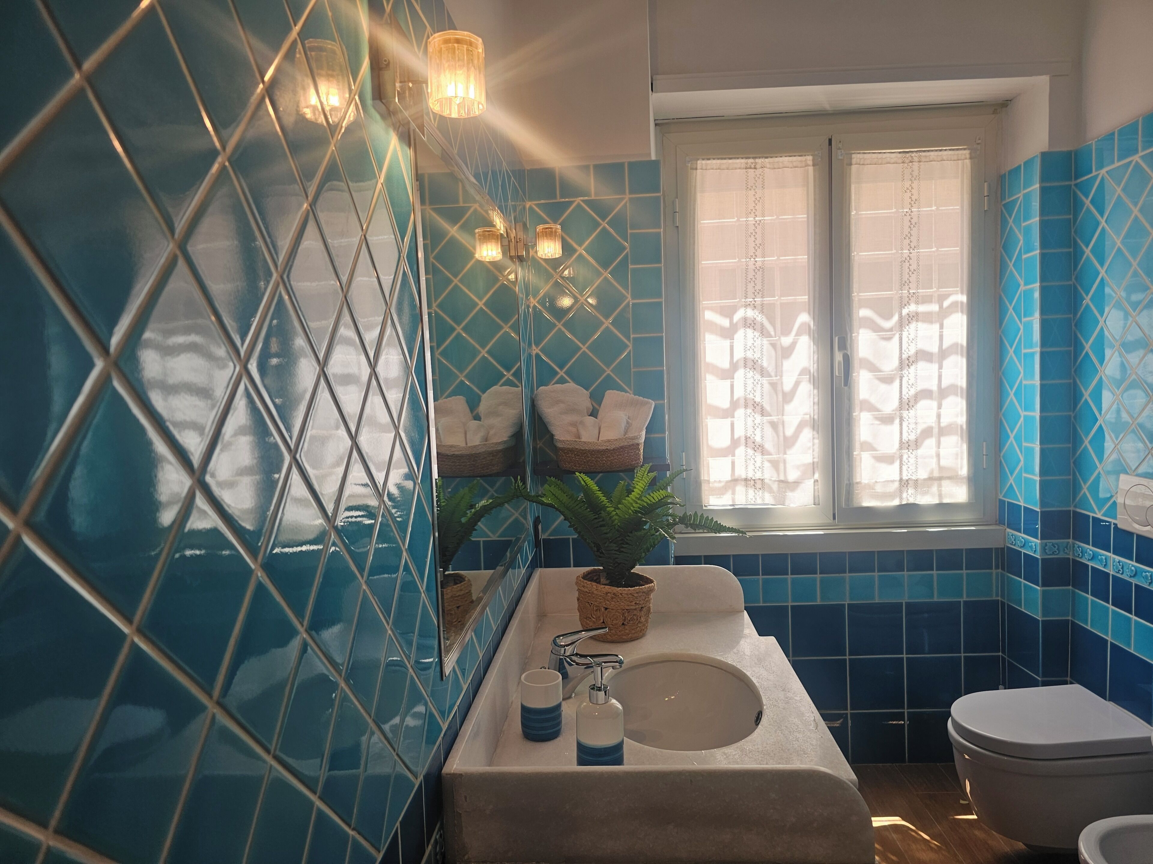 Ein Badezimmer mit blauen Fliesen, einem Waschbecken, einer Toilette und einem Fenster mit Vorhängen.