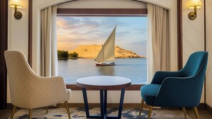A room with two chairs and a table, a large window with curtains, and a view of a sailboat on water.