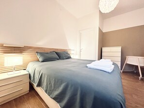 Una cama bien hecha con cubrecama y almohadas grises, una mesita de noche con lámpara, un armario blanco y un escritorio pequeño con silla.
