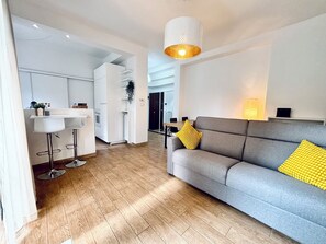 Un salón moderno con un sofá gris, almohadas amarillas, un taburete blanco y una zona de cocina con armarios blancos y una encimera blanca.