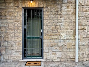 Fachada de pared de piedra con una puerta de metal, un tapete con la palabra 'WELCOME' y una lámpara sobre la puerta.