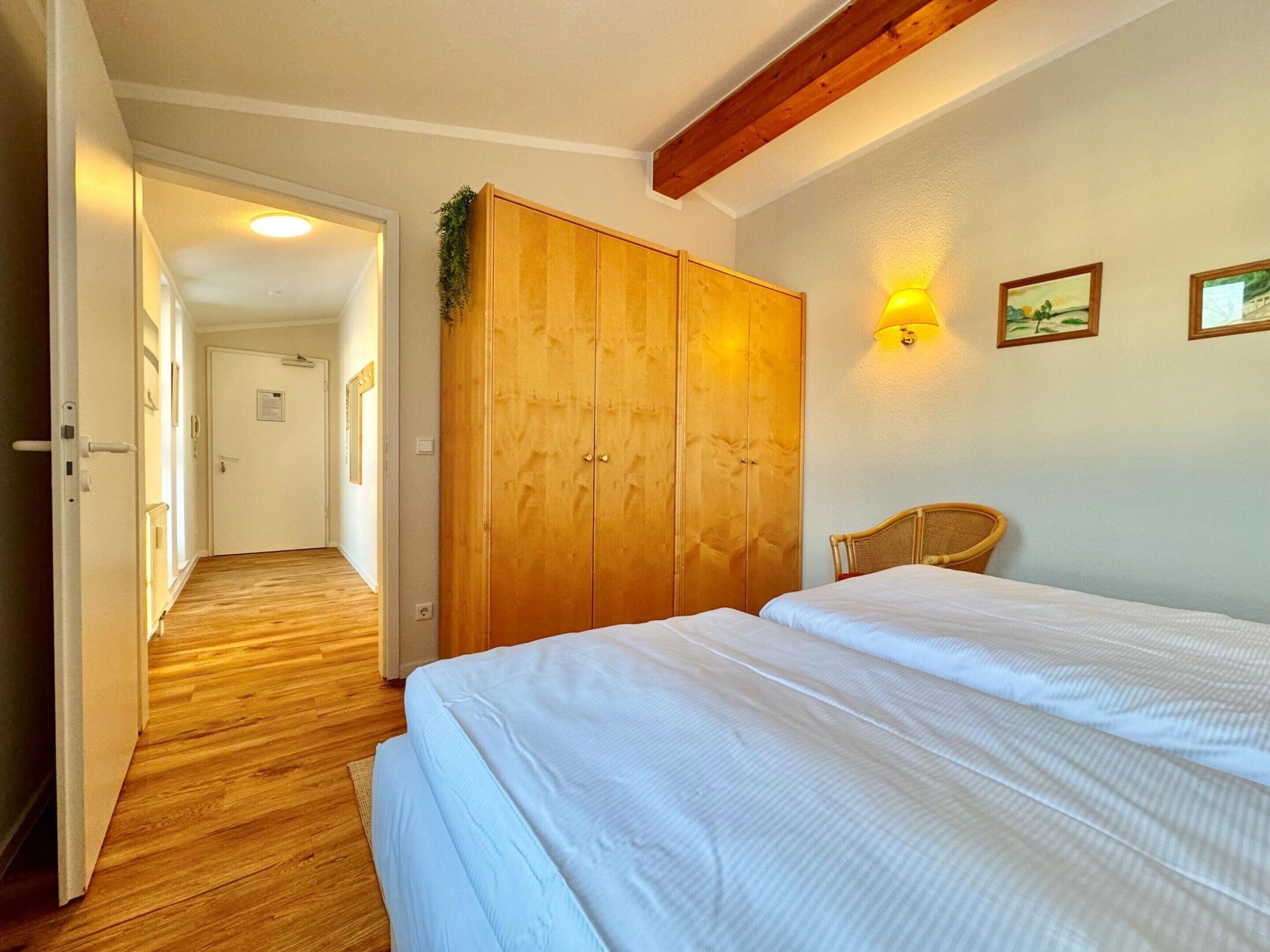 Een slaapkamer met een houten garderobe, een bed met wit beddengoed, een stoel en een gang naar een andere kamer.