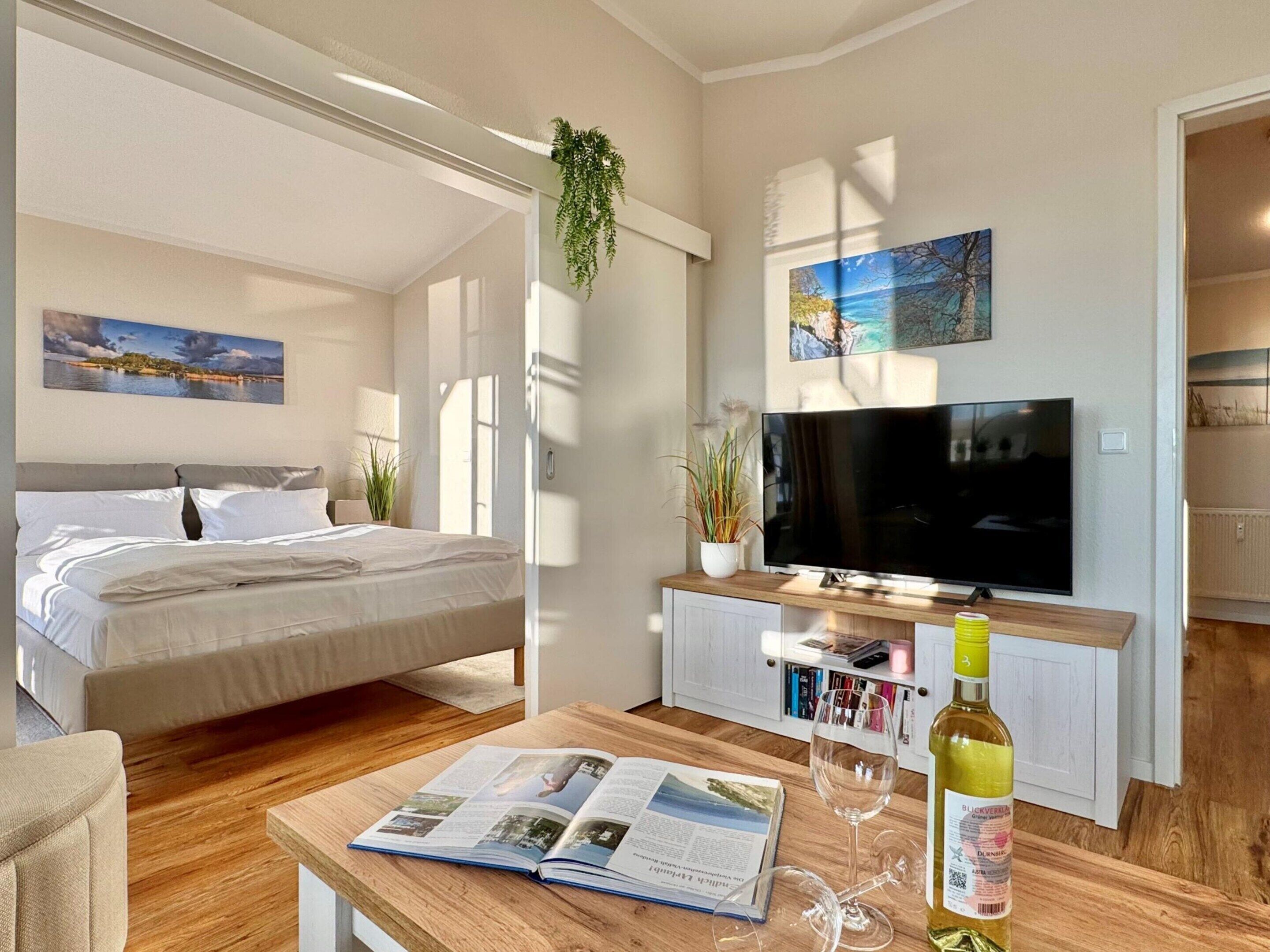 A modern hotel room with a bed, a TV, a wooden coffee table with a magazine, a wine glass, and a bottle of wine.