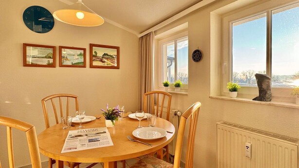Een eetgedeelte met een ronde tafel, stoelen, een klok, ingelijste foto’s, een raam met uitzicht en een radiator.