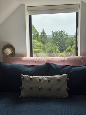 A bedroom with a view of a forest through a window, a bed with dark blue bedding, and a decorative pillow with pom-poms.