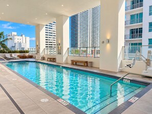 Una piscina con indicador de profundidad de 3 pies, rodeada de sillas de descanso y un banco, con vista a edificios altos.