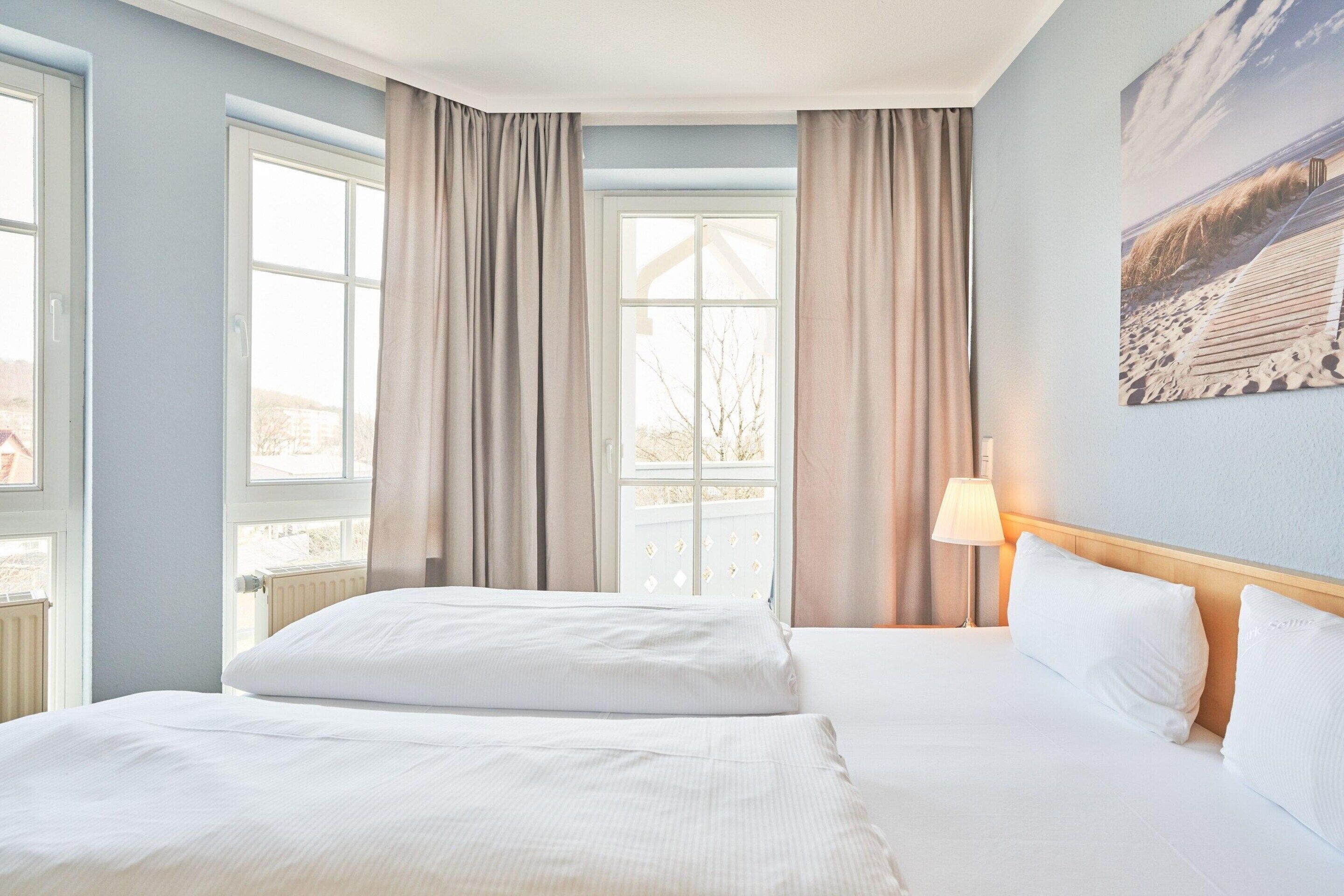 A hotel room with two beds, large windows with curtains, a lamp, and a painting on the wall.