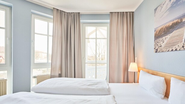 Ett hotellrum med två sängar, stora fönster med gardiner, en lampa och en tavla på väggen.