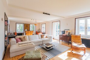 A spacious living room with a large sofa, a coffee table, and a chair. There is a bookshelf, a radiator, and a window with a view of trees.