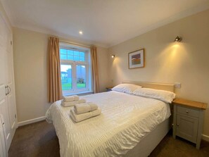 A bedroom with a bed, a nightstand, a window with curtains, and a wall-mounted light. - Atlantic 25 | Contemporary 4BR Holiday Pad Newquay (Newquay)