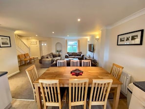 A dining area with a wooden table and chairs, a staircase leading to another room, and a living area with a sofa and a TV. - Atlantic 25 | Contemporary 4BR Holiday Pad Newquay (Newquay)