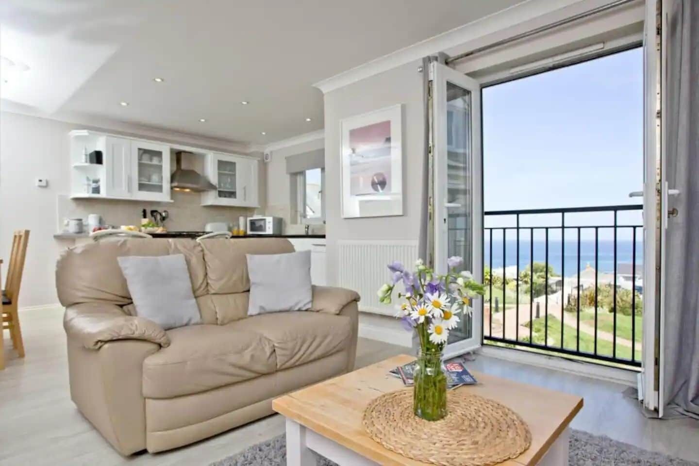 Ein Wohnzimmer mit einem beigefarbenen Sofa, einem Holztisch mit einer Vase, einer Küche im Hintergrund und einem Balkon mit Meerblick.