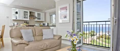 Ein Wohnzimmer mit einem beigefarbenen Sofa, einem Holztisch mit einer Vase, einer Küche im Hintergrund und einem Balkon mit Meerblick.
