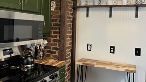 Une cuisine avec des meubles de couleur verte, un mur en briques, une cuisinière en acier inoxydable et une étagère en bois où sont posées des tasses.