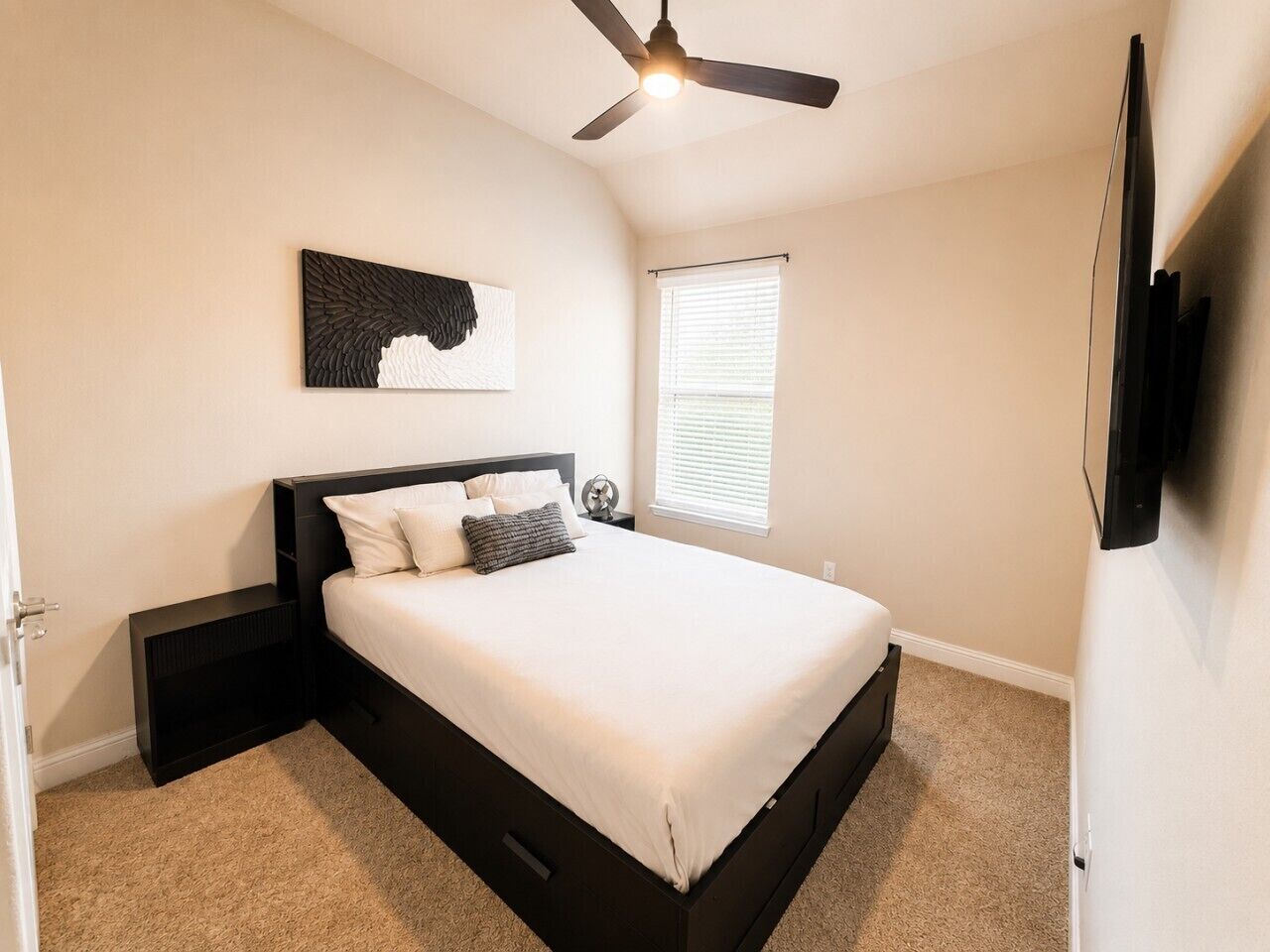 A bedroom with a bed, a ceiling fan, a window with blinds, and a wall-mounted TV.