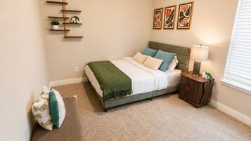 Quarto com cama, mesa de cabeceira, luminária, decoração na parede e uma pequena planta.