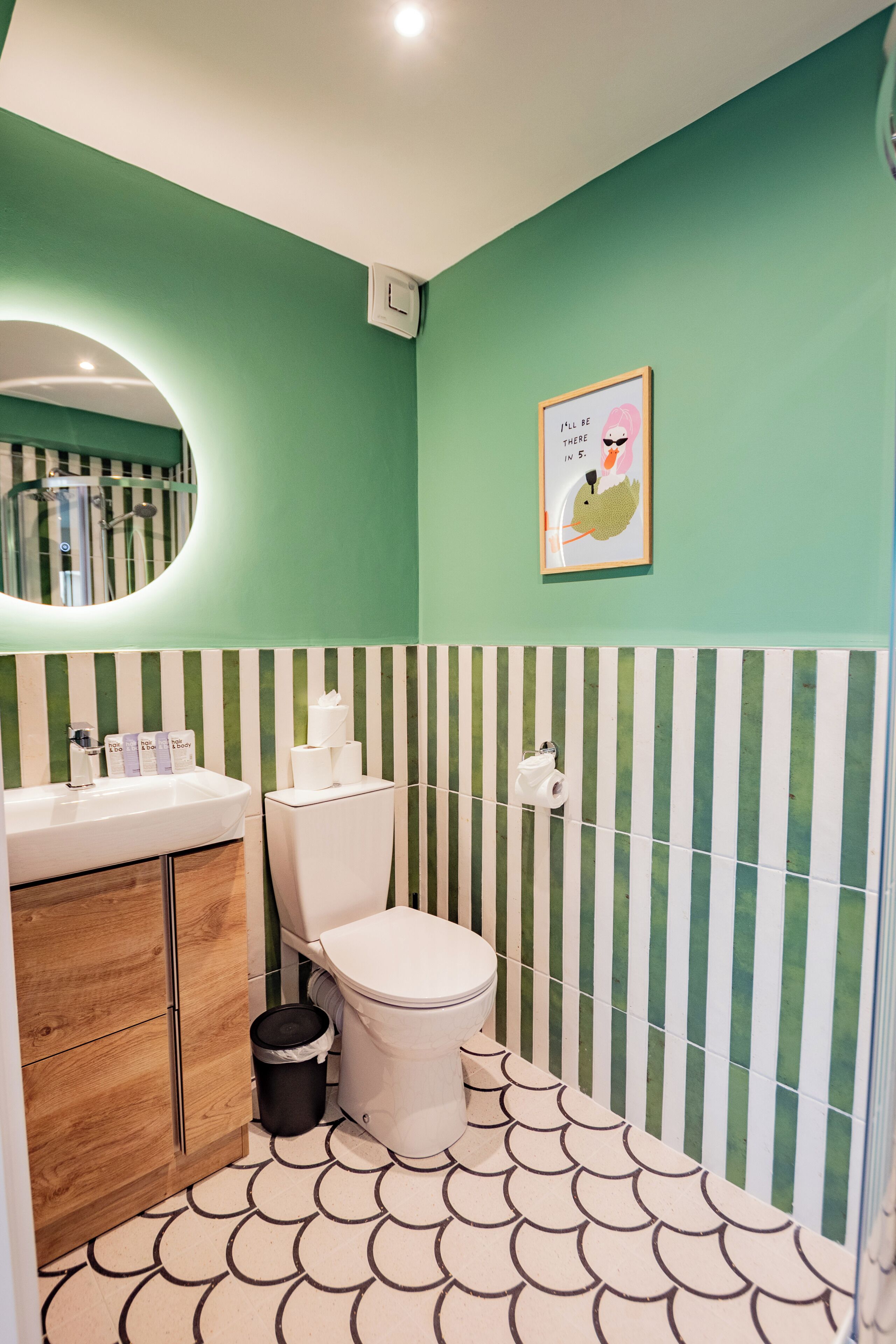 A bathroom with a green wall, striped wallpaper, a toilet, a sink, a mirror, and a framed picture.