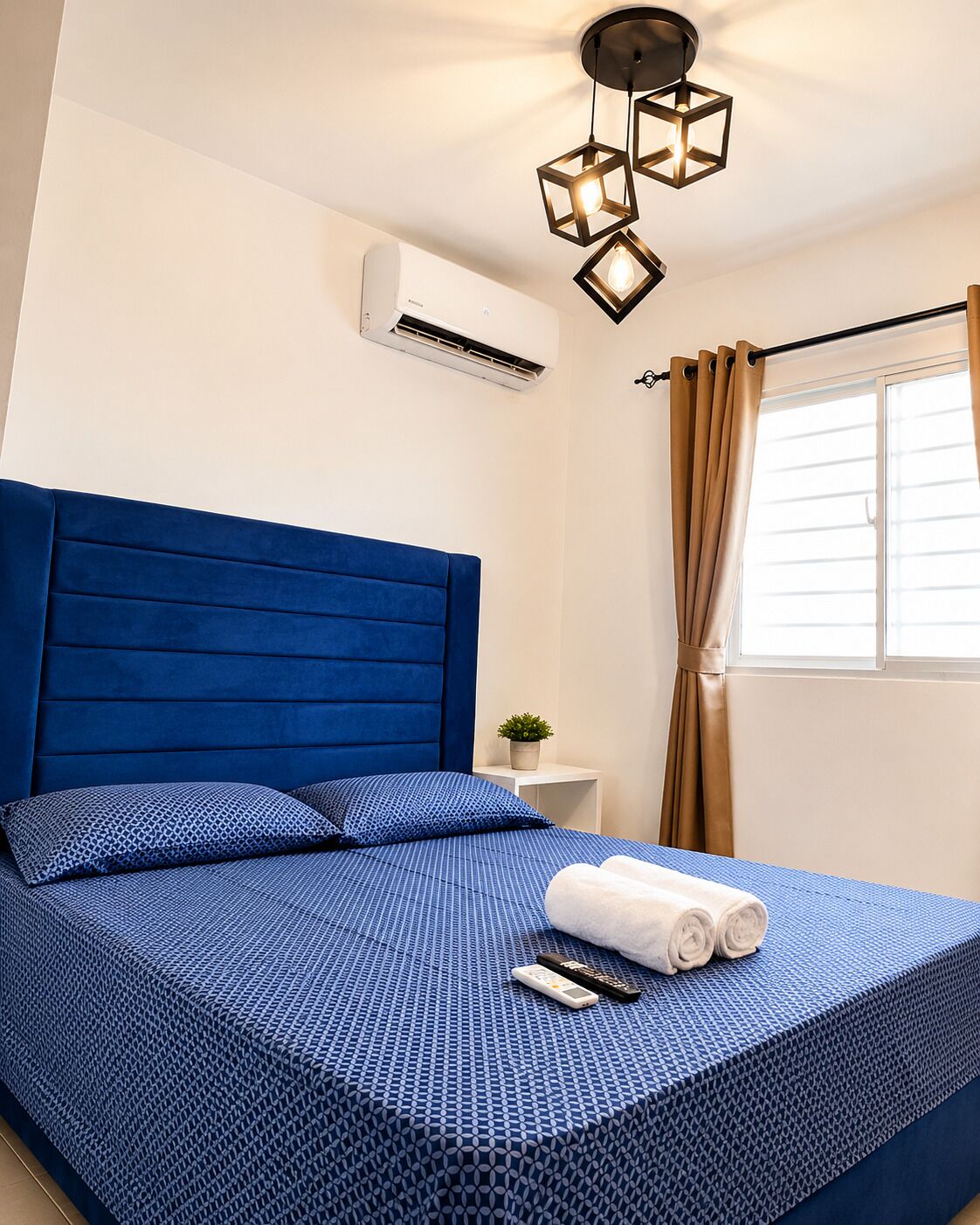 A hotel room with a blue bed, a small bedside table with a plant, and a window with curtains.