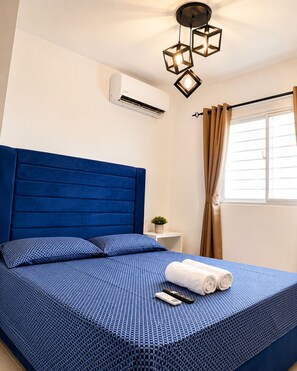Ein Hotelzimmer mit einem blauen Bett, einem kleinen Nachttisch mit Pflanze und einem Fenster mit Vorhängen.
