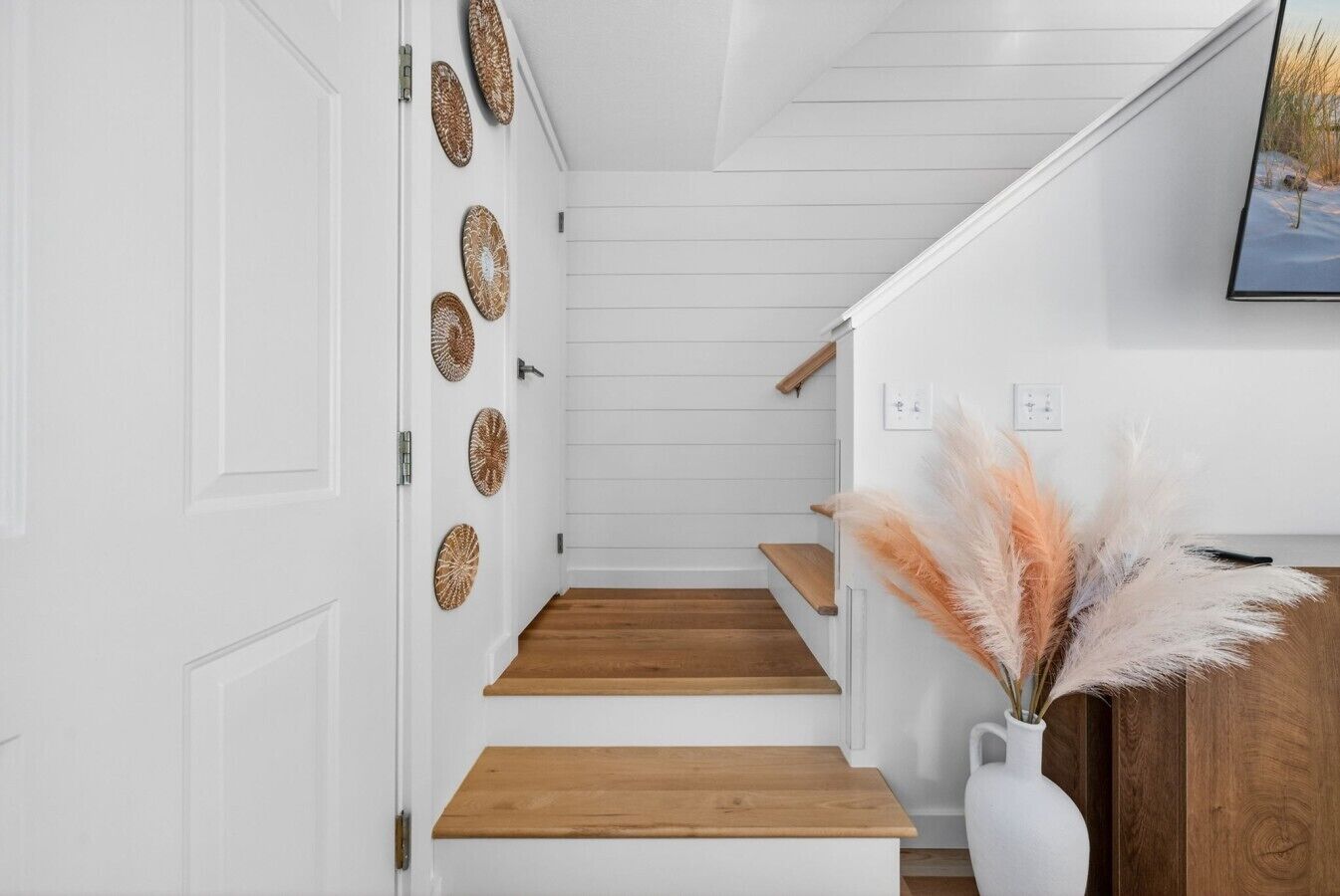 Eine Treppe mit Holzdielen und weißen Wänden, eine weiße Tür mit dekorativen Platten und eine Vase mit Pampasgras.
