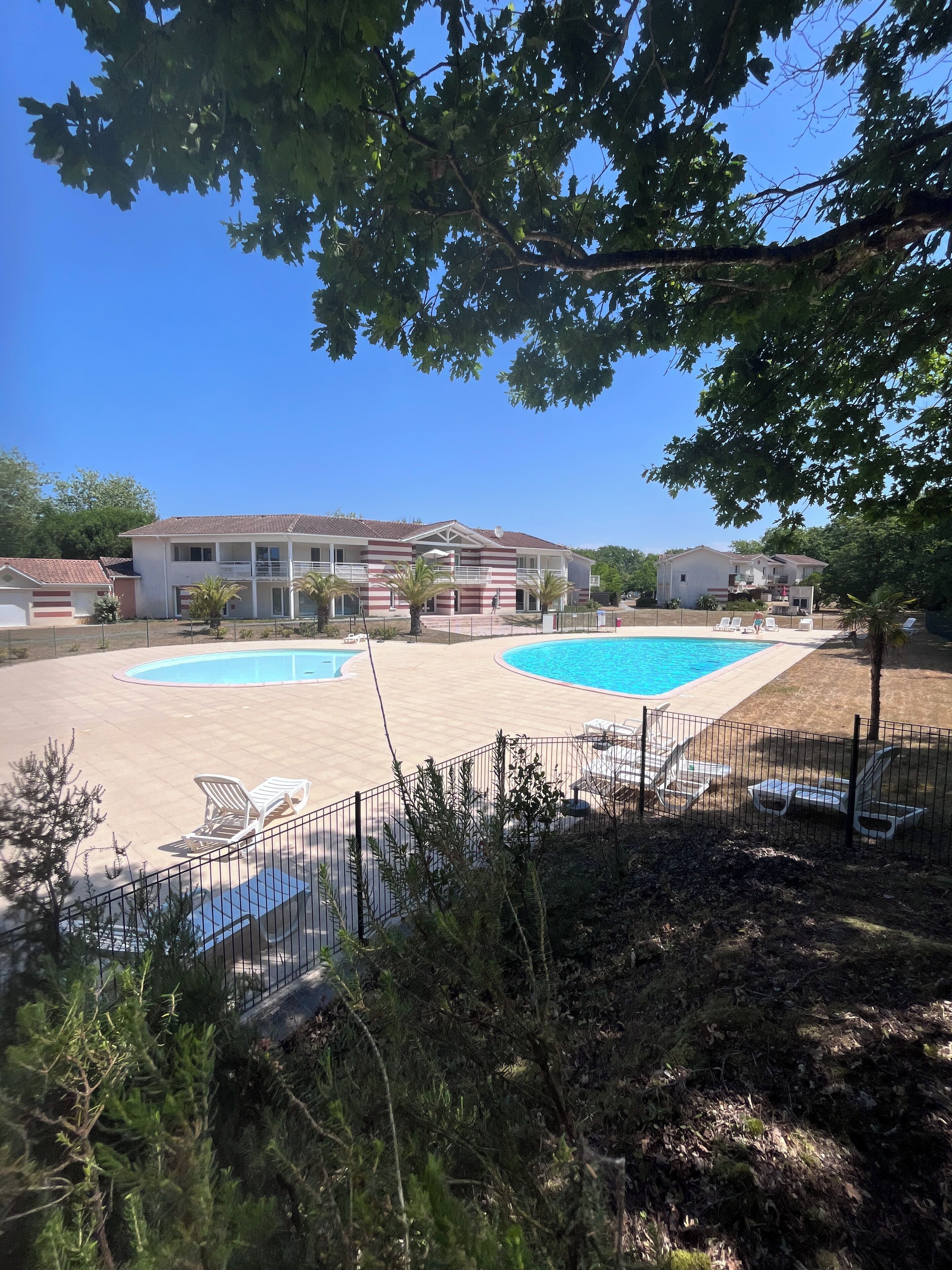 Ein Wohngebiet mit zwei Schwimmbecken, einer umzäunten Terrasse mit Liegestühlen und einem Gebäude mit einem Ziegeldach.