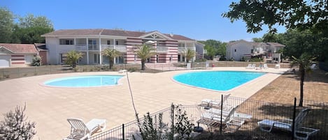 A residential area with two swimming pools, a fenced patio area with lounge chairs, and a building with a tiled roof.
