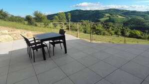 Een terras met een tafel en stoelen, een metalen hekwerk en uitzicht op glooiende heuvels.