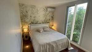Une chambre à coucher comprenant un lit, deux lampes de chevet, un climatiseur fixé au mur et une grande fenêtre donnant sur un paysage verdoyant.