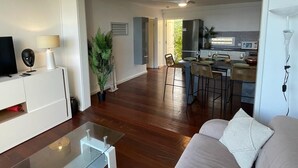 Un salon moderne avec une table basse en verre, un canapé et un téléviseur posé sur un meuble blanc. On aperçoit également un coin repas avec une cuisine en arrière-plan.