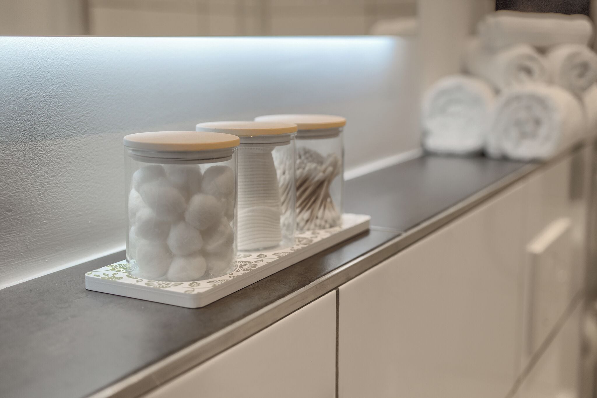 Three glass jars with wooden lids on a countertop, a rolled towel, and a white decorative tray.