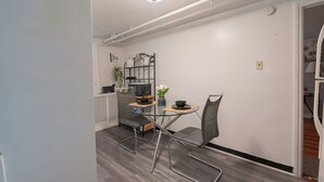Een compacte leefruimte met een eethoek met een glazen tafel en metalen stoelen, een keuken met een magnetron en koffiezetapparaat, en een zichtbaar plafond met blootliggende leidingen.