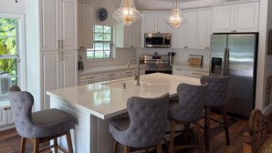 Una cocina moderna con armarios blancos, electrodomésticos de acero inoxidable, una isla central con taburetes y luminarias colgantes, además de un ventilador de techo.