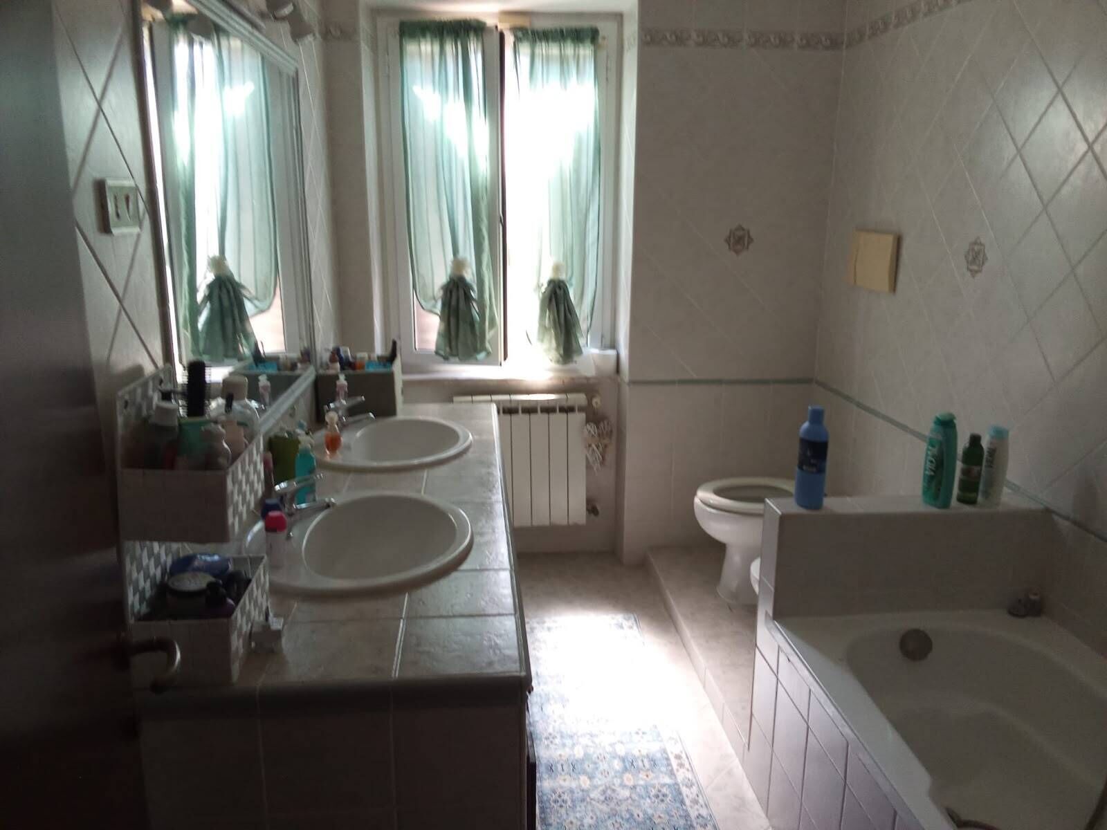 Une salle de bain avec deux lavabos, des toilettes, une baignoire et une fenêtre avec des rideaux.