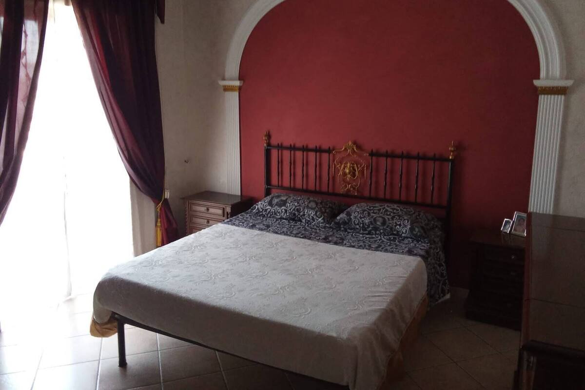 Una camera da letto con un letto, un comodino, una cassettiera e un muro rosso con una finestra ad arco.