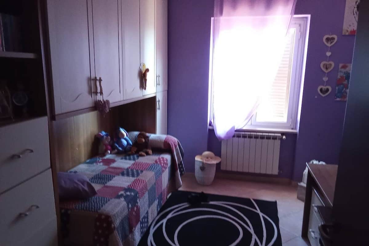Una camera da letto con un letto, un termosifone, una finestra con tende, un cestino e un comò.