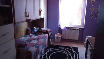 Quarto com cama, radiador, janela com cortinas, lixeira e cômoda.