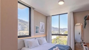 Ein Hotelzimmer mit einem Bett, zwei Fenstern, einem Spiegel und Blick auf eine Berglandschaft.
