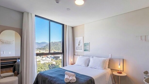 A hotel room with a large window, a bed with white linens, a bedside table with a lamp, and a view of the landscape. - Modern Apt. W/city View in Urubici Ust0409 (Urubici)