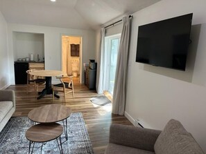 Una sala de estar con sofá, una mesa de centro, un televisor montado en la pared y un comedor con mesa y sillas.