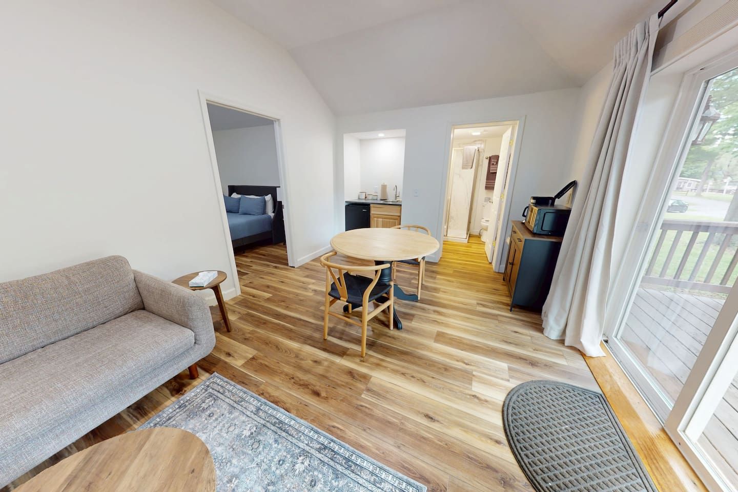Un espacio habitable compacto con sofá, una mesa redonda y una cocina visible a través de una puerta abierta.