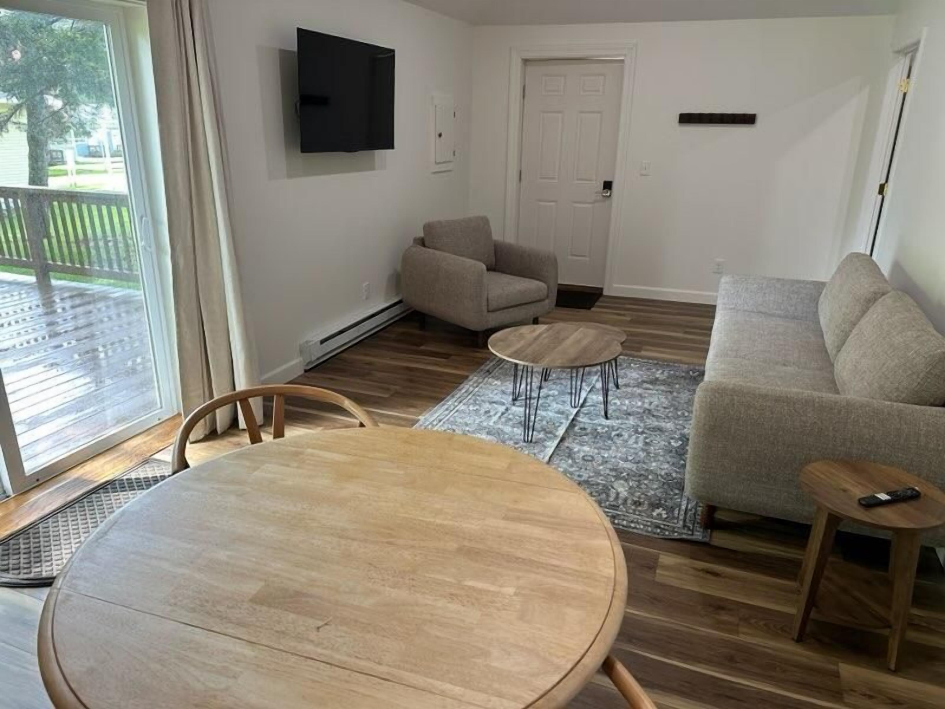 Una sala de estar con una mesa de comedor de madera, dos sofás, un televisor montado en la pared y un ventanal corredizo que da a un área exterior.