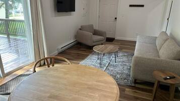 Una sala de estar con una mesa de comedor de madera, dos sofás, un televisor montado en la pared y un ventanal corredizo que da a un área exterior.