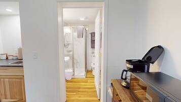 Un couloir étroit mène à une salle de bain équipée d’une douche, de toilettes et d’un lavabo. Une machine à café est posée sur un comptoir en bois.