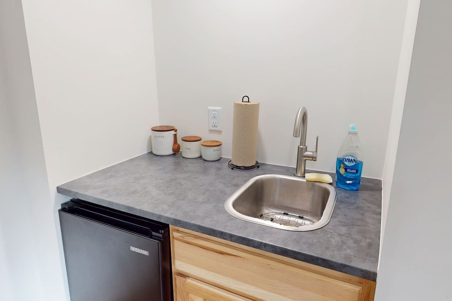 Cocina compacta con lavavajillas, un lavamanos con grifo y frascos para café, té y azúcar.