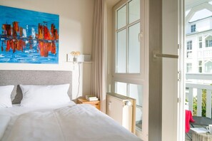 A hotel room with a bed, a painting, a nightstand, a lamp, and a window with a view of a balcony. - Moderne Ferienwohnung in Sellin (Sellin)