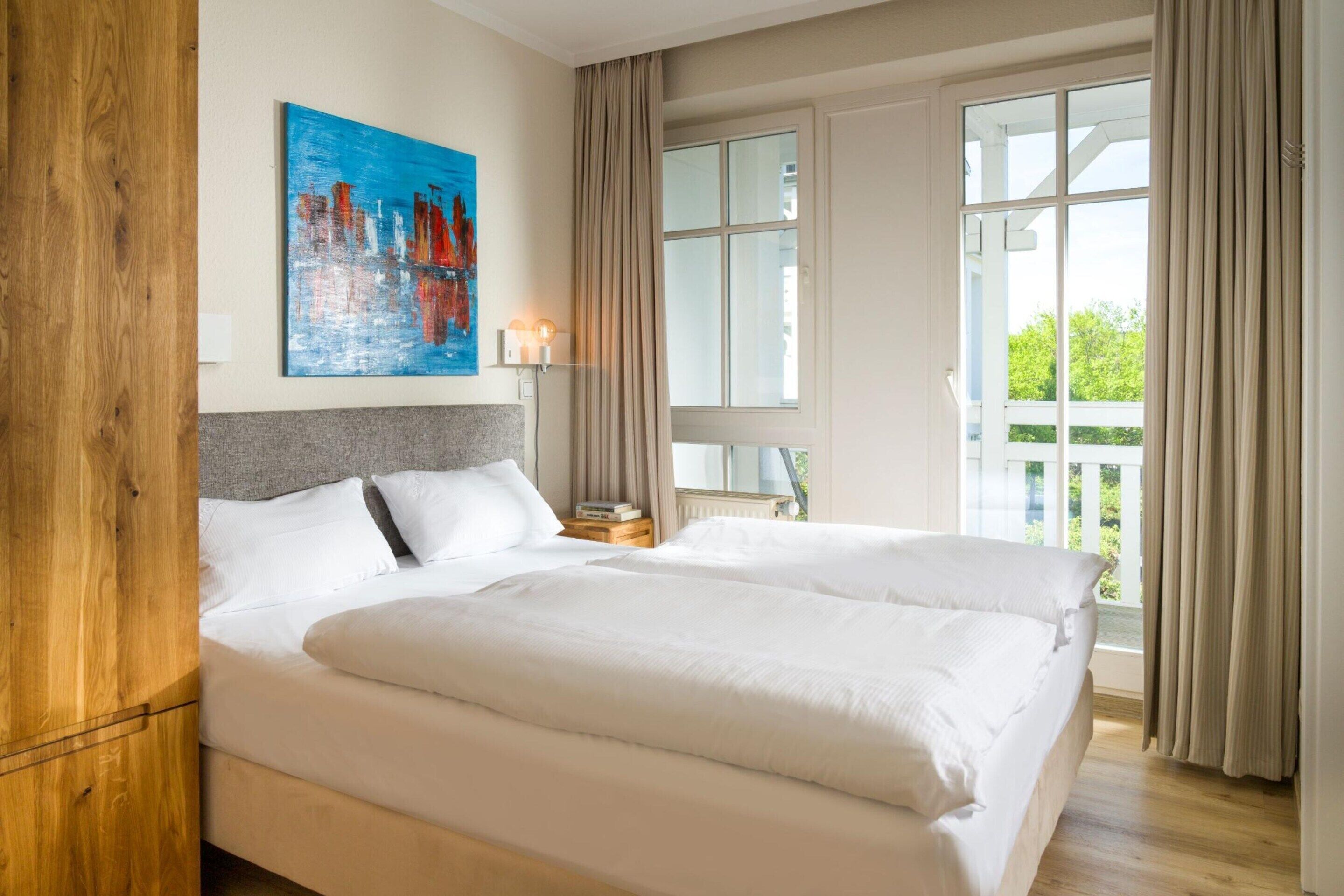 Un lit double avec du linge de lit blanc, une tête de lit en bois, un tableau au mur, une table de chevet avec une lampe et une fenêtre avec des rideaux.
