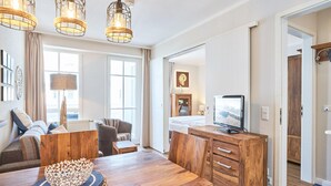 Ein modernes Wohnzimmer mit einem Holzesstisch, einer Couch und einem Fernseher. Es gibt drei Pendelleuchten, eine Holzkommode und ein großes Fenster.
