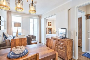Ein modernes Wohnzimmer mit einem Holzesstisch, einer Couch und einem Fernseher. Es gibt drei Pendelleuchten, eine Holzkommode und ein großes Fenster.