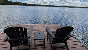 A wooden dock with two Adirondack chairs and a picnic table, overlooking a calm lake with a forested shoreline under a partly cloudy sky. - Beautiful 3 bed/2bath updated cabin w sandy swimmable frontage & great fishing (Eagle River)
